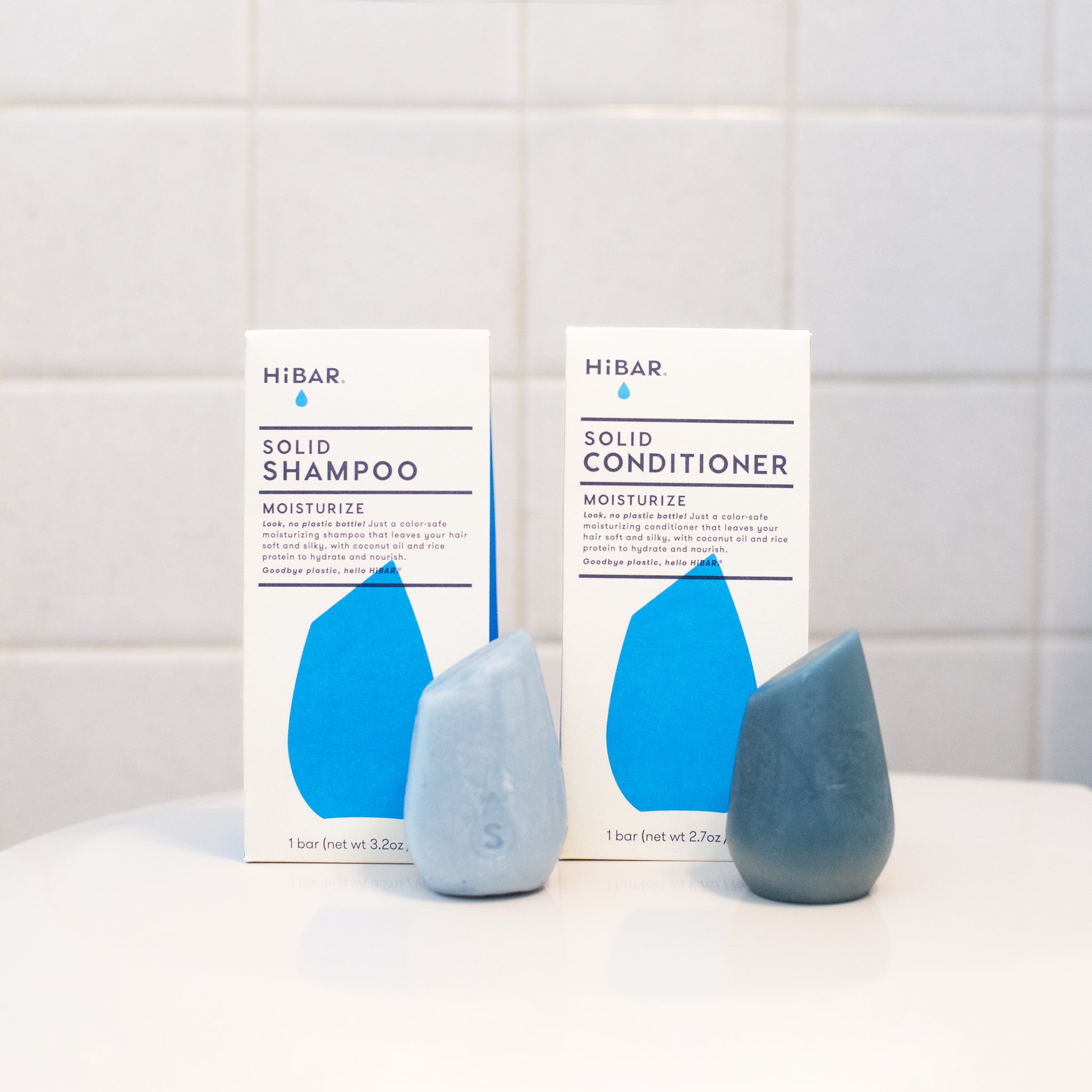 Two HiBar MOISTURIZE solid hair care products: a light blue-gray shampoo bar next to a deep-blue conditioner bar, showcasing their plastic-free design, coconut oil, and rice protein ingredients for soft, silky hair.