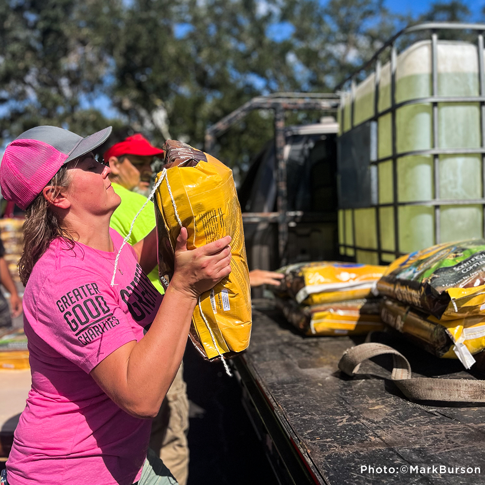 A woman in a pink Greater Good Charities shirt lifts a heavy bag of supplies from a truck during disaster relief efforts. Photo by Mark Burson.