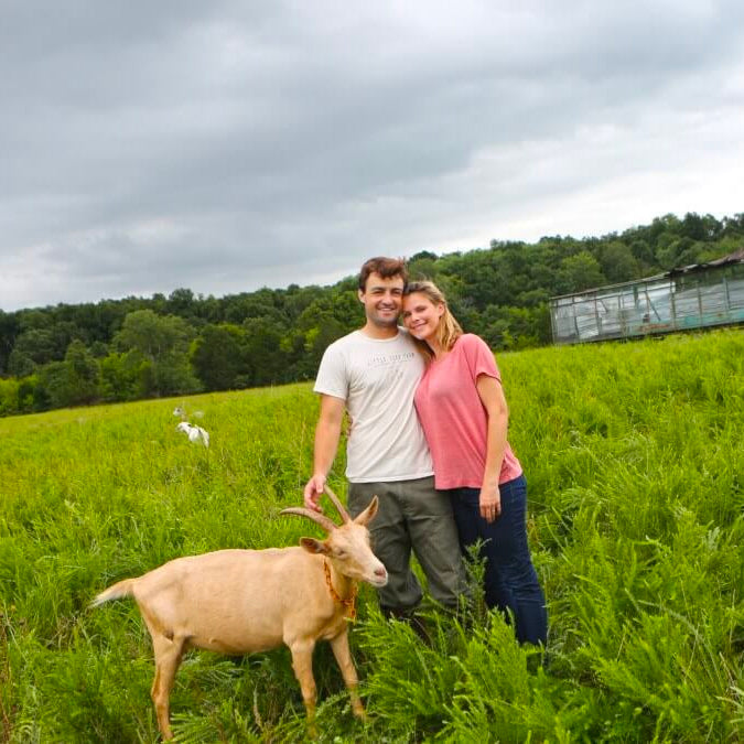 A couple stands in a lush green field on a cloudy day, a light brown goat next to them.