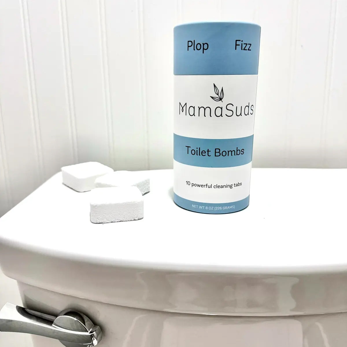 A close-up of white toilet cleaning tabs scattered beside the MamaSuds pouch, emphasizing their compact, fizzy form made with vegan, non-toxic ingredients.