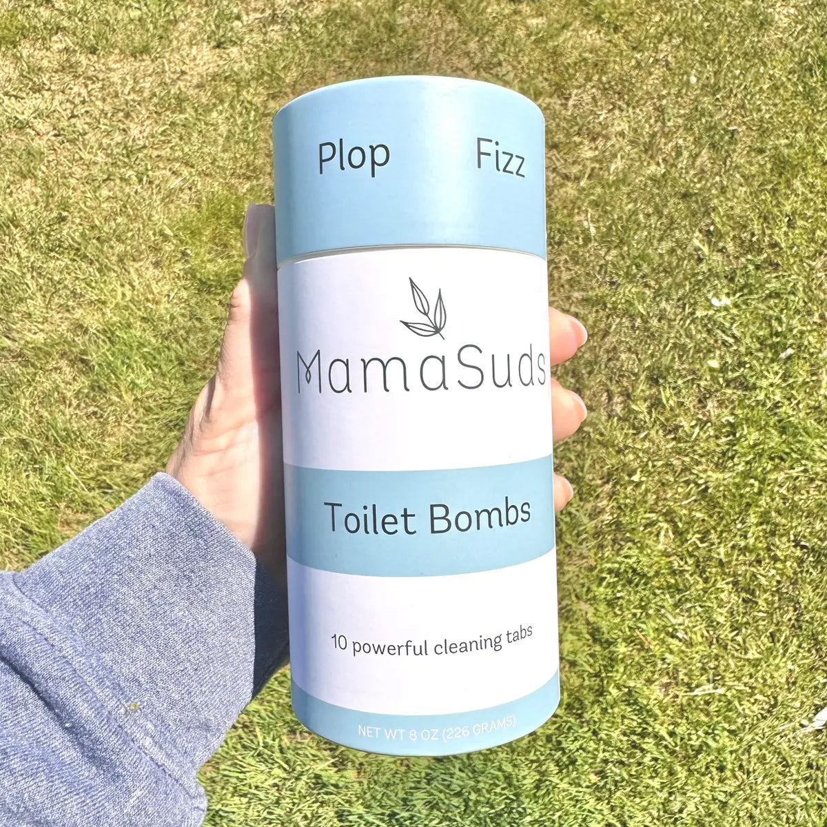 An overhead view of a hand holdong the package of MamaSuds Toilet Bombs, in a green grass background.