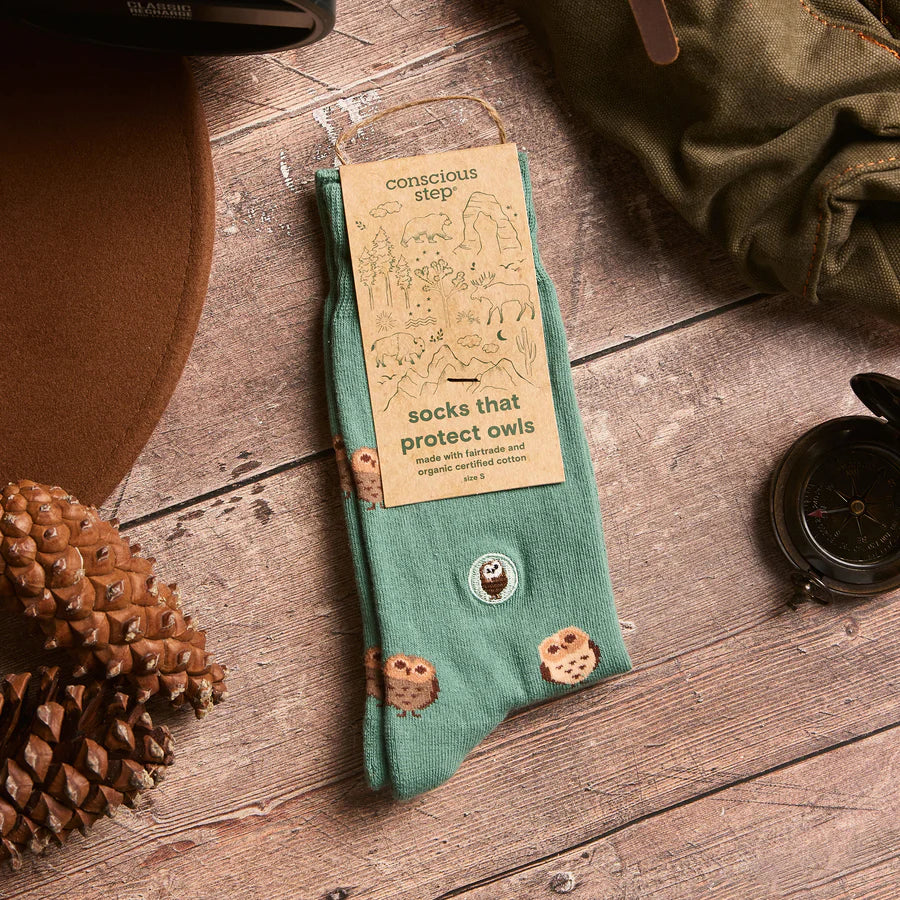 A pair of green socks with a barn owl pattern is shown in its packaging. The tag identifies them as "socks that protect owls," made with fairtrade and organic certified cotton. The socks are set against a wooden background with a pinecone, hat, and compass.