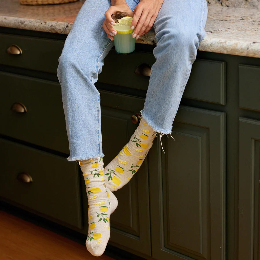 A person is shown wearing light-colored socks with a yellow lemon and green leaf pattern and torn-hem jeans. The person is sitting on a countertop, holding a glass with a yellow liquid.
