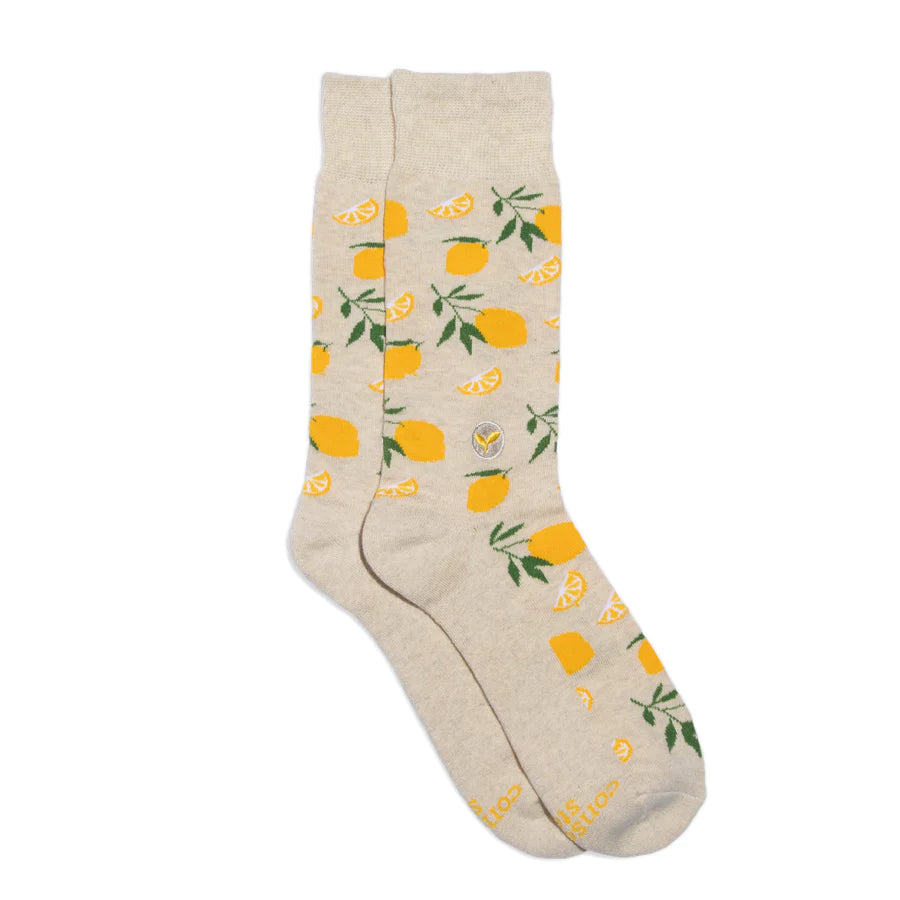 A pair of light-colored Conscious Step socks with a pattern of yellow lemons and green leaves is shown against a white background. The socks are lying flat, and the brand name is visible on the bottom of the foot.