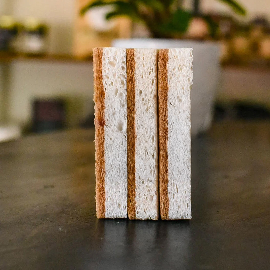 Three eco-conscious natural sponges with labels stacked on a kitchen countertop, emphasizing their biodegradable and non-abrasive qualities.