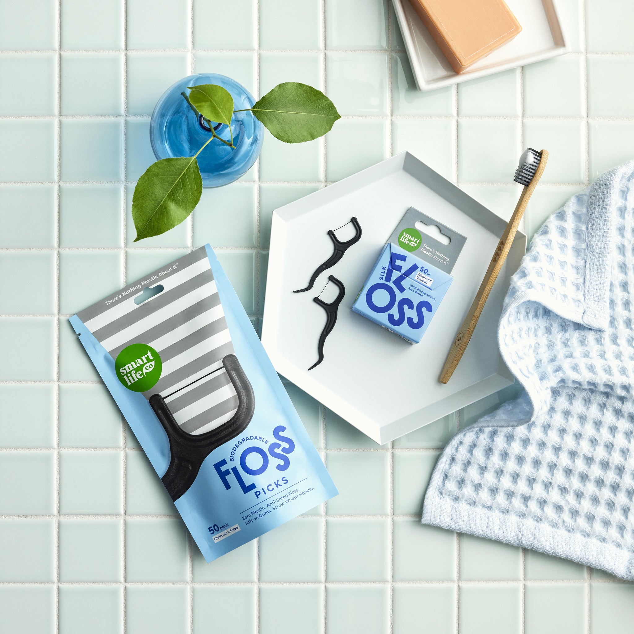 A cute overhead shot of charcoal-infused floss picks on a tray with a bamboo toothbrush and a blue towel, showcasing the product in a clean, modern bathroom setting.
