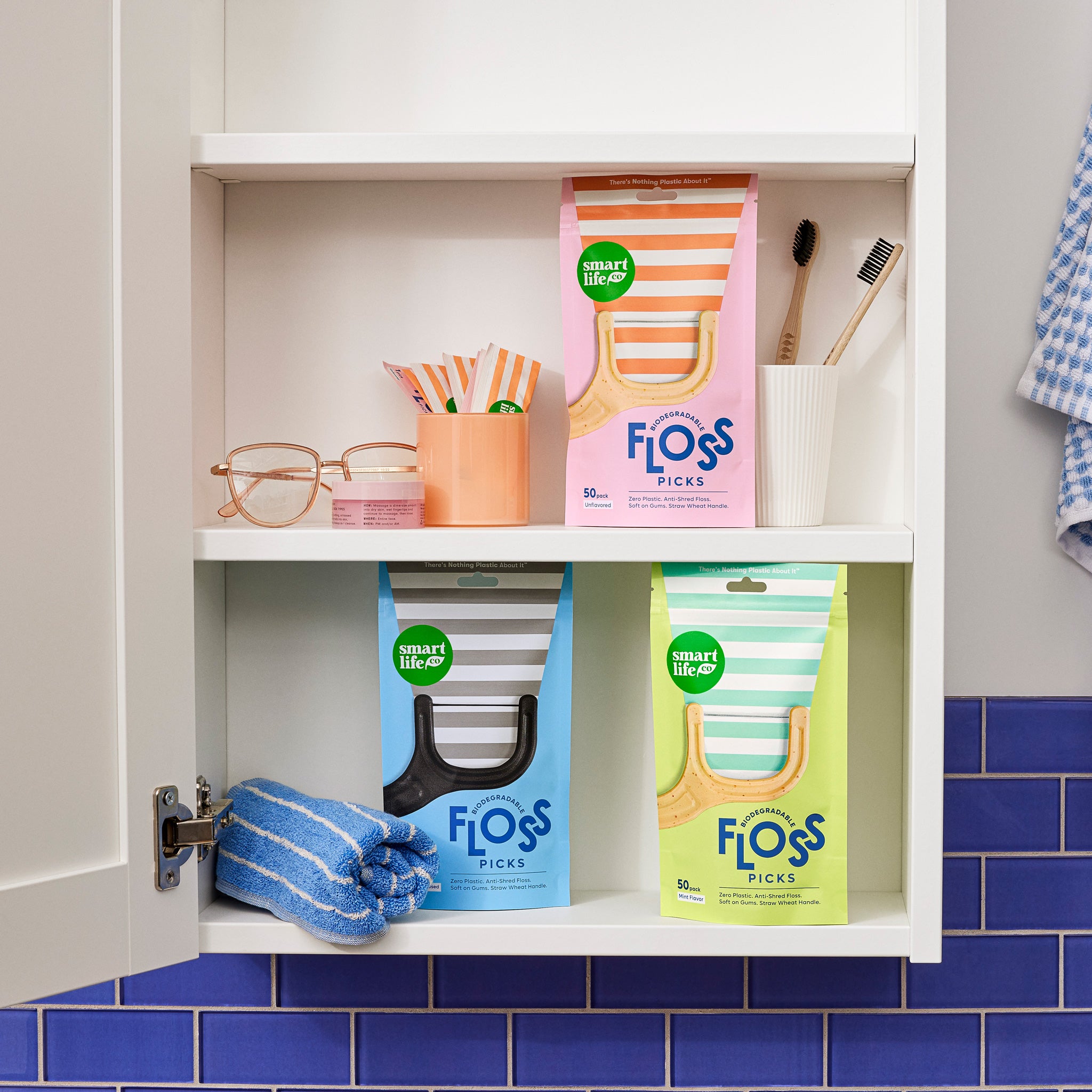 A cute bathroom medicine cabinet is neatly organized with Smart Life Co. floss pick boxes and two bamboo toothbrushes. The blue, pink, and green boxes are arranged on the shelves, showing the products in a home setting.