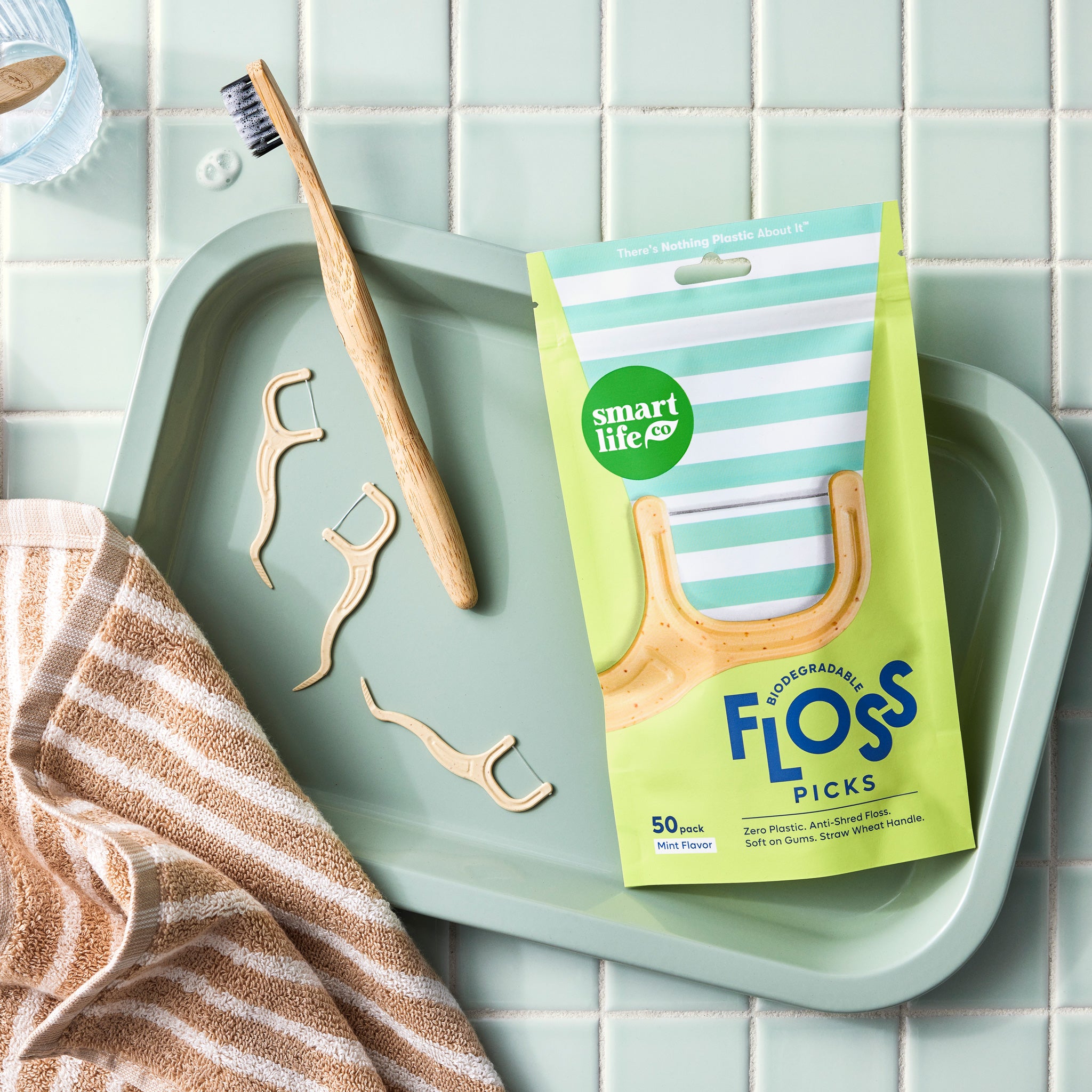 A cute overhead shot of mint-flavored floss picks pouch on a tray with a bamboo toothbrush and a beige towel, showcasing the product in a clean and modern bathroom setting.