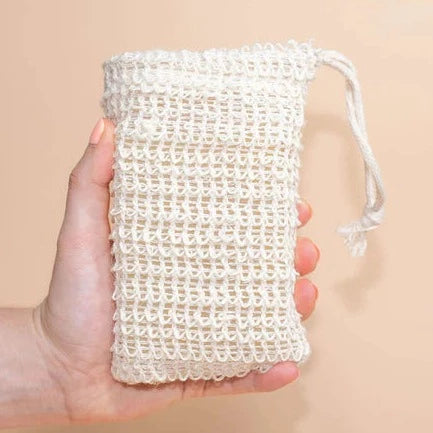 A hand holds a textured, light beige, reusable soap bag made from a woven material. The bag's drawstring is visible at the top