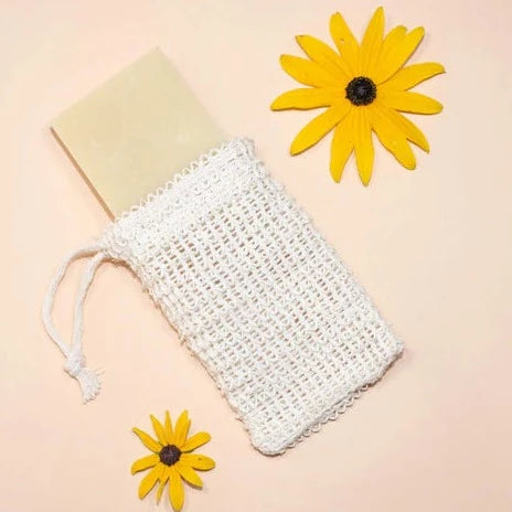 A light-colored bar of soap is half-inserted into a beige, woven soap saver bag. Two yellow, daisy-like flowers are also on the pinkish-beige background.