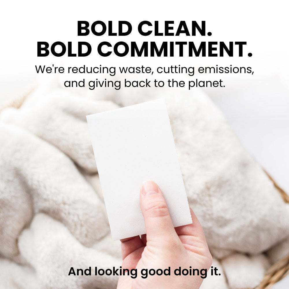 A hand is holding a piece of paper with bold text that reads "BOLD CLEAN. BOLD COMMITMENT. We're reducing waste, cutting emissions, and giving back to the planet. And looking good doing it." The background appears to be neutral, focusing on the hand and paper.
