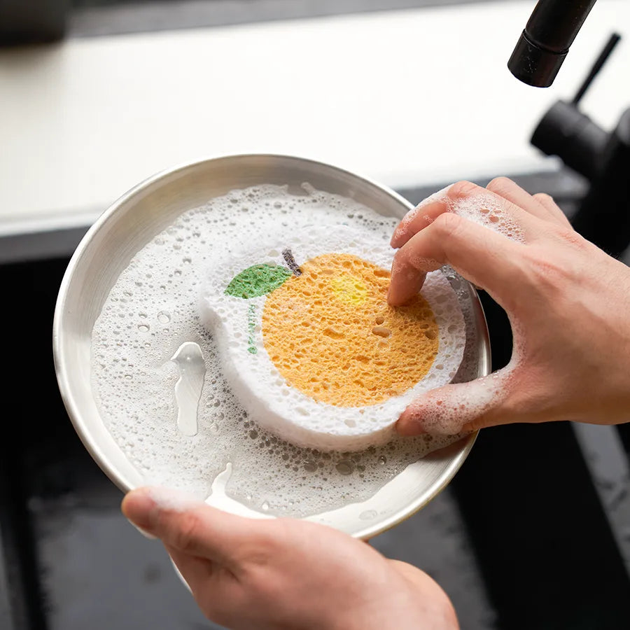 A person is using a PLANTISH orange-shaped sponge to wash a silver bowl. The porous sponge, which has a yellow-orange orange slice and green leaf design is covered in soap bubbles.