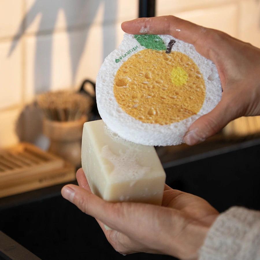 A person is using a PLANTISH orange-shaped sponge to lather a bar of light-colored soap. The sponge is porous and features a yellow-orange orange slice and green leaf design on a white background.