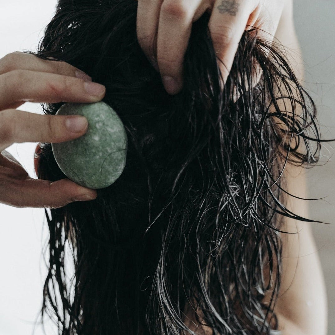 A speckled green shampoo or conditioner bar is held by a hand and applied directly to wet dark hair, lathering the strands.