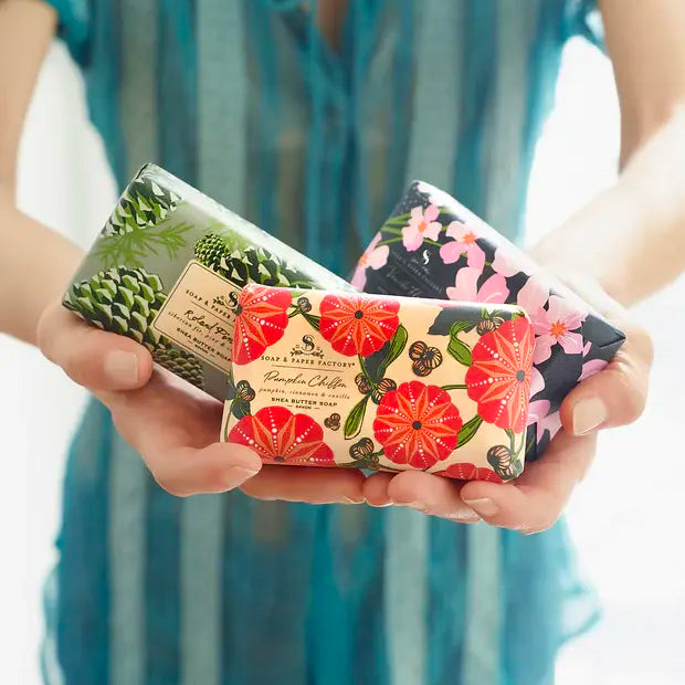 A person holds three rectangular 'Soap & Paper Factory' shea butter soaps. Each soap is wrapped in a different, colorful floral or botanical paper. Each features a different scent.