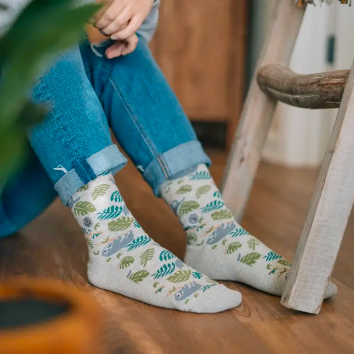 A person is shown wearing cuffed blue jeans and gray socks with a pattern of sloths and leaves. The person is sitting on a wood floor next to a plant and a wooden ladder.