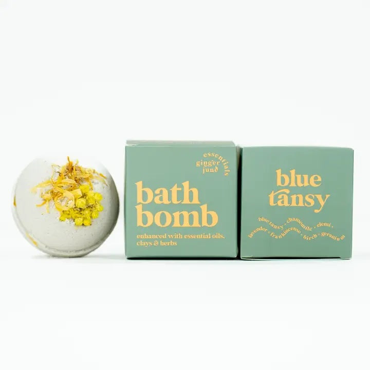 A light-colored bath bomb is shown next to two light green boxes. The bath bomb is topped with dried yellow flowers. The boxes are labeled ginger june essentials and blue tansy with the essential oils blue tansy, chamomile, lavender, elemi, frankincense, birch and geranium listed.