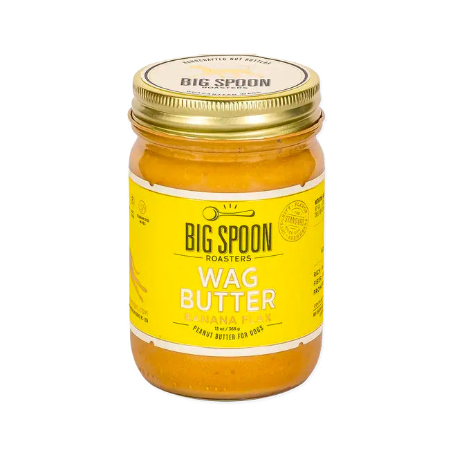 A glass jar with a gold lid and a yellow label is for "Big Spoon Roasters Wag Butter Banana Flax," a peanut butter for dogs. The jar contains a yellow-orange butter.