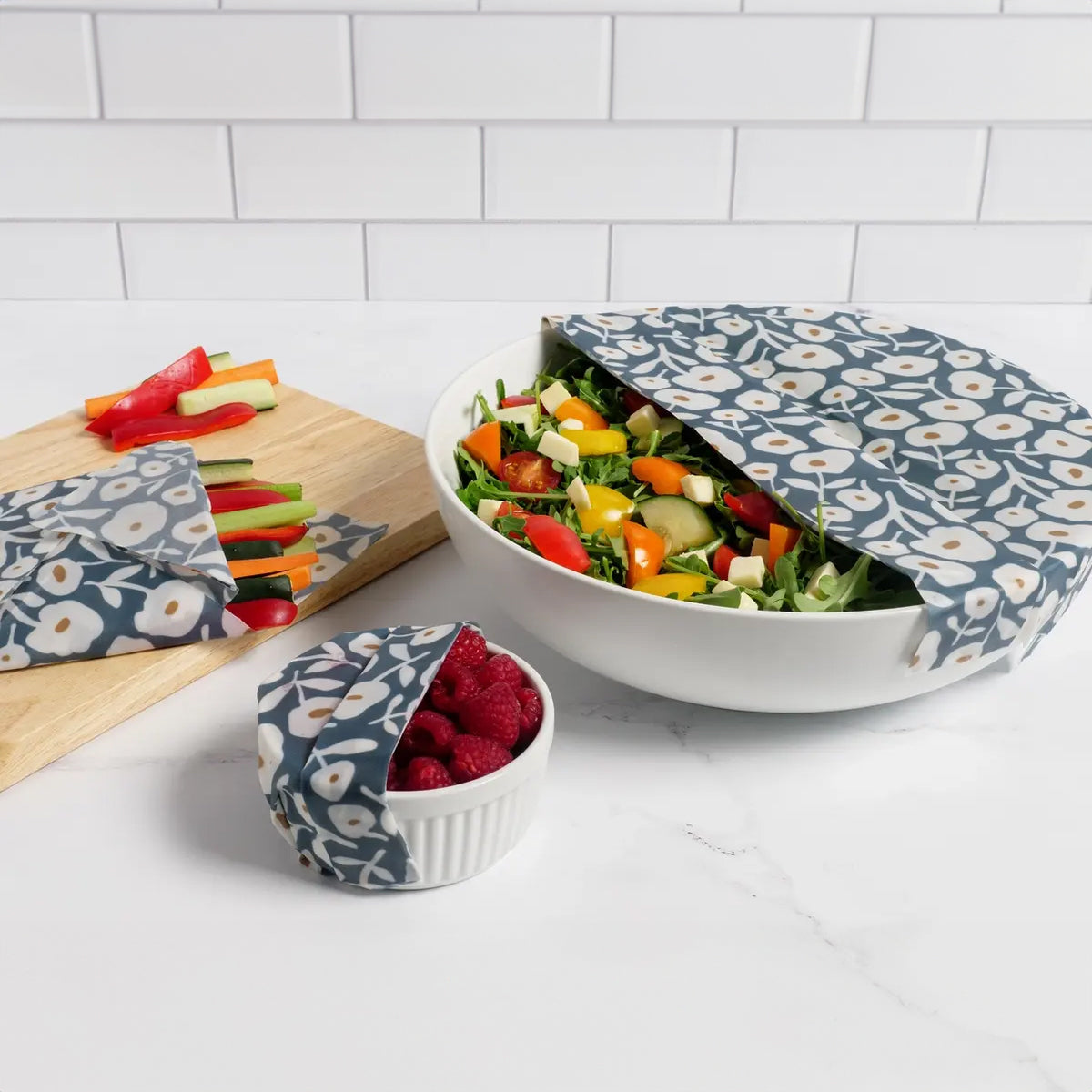 A bowl of salad wrapped in Reusable Beeswax Food Wrap placed next to a bowl of fresh vegetables on a table also wrapped.