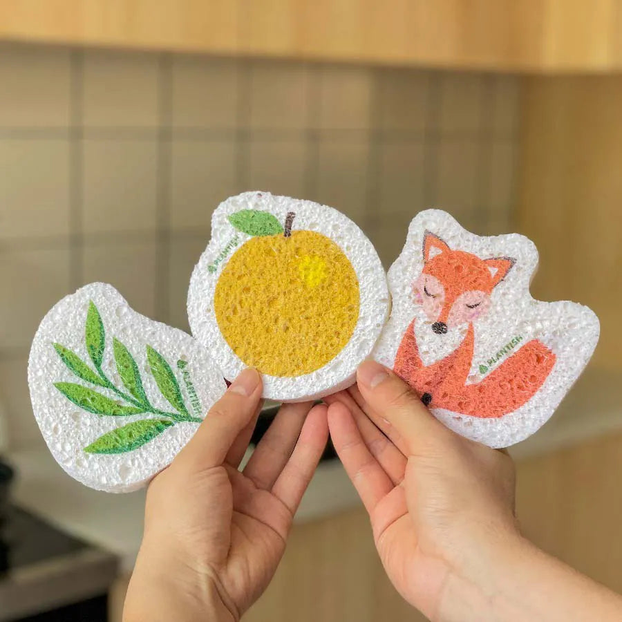 A person's hands hold three expanded PLANTISH pop-up sponges in a fan-like arrangement. The sponges are shaped like a green leaf, an orange and a fox.