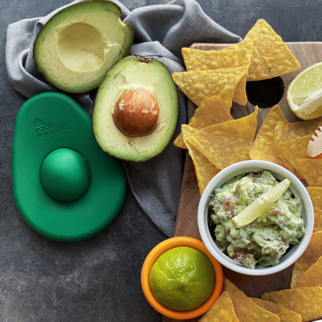 A delightful scene of sustainable food storage, where a reusable silicone avocado hugger keeps a fresh avocado half from browning. A bright orange food saver is also shown covering a lime, all arranged with a bowl of delicipus guacamole and nachos.