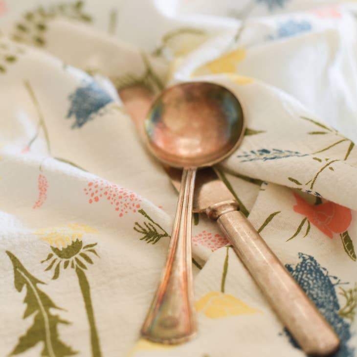 A charming white kitchen towel with a whimsical pattern of pink, yellow and teal wildflowers is partially covered by a vintage spoon and knife.