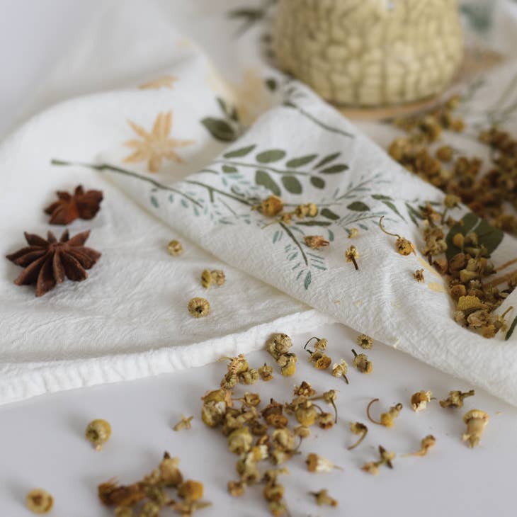 A white kitchen towel with a light green leaf and beige botanical pattern is folded with scattered dried chamomile flowers and star anise pods nearby.