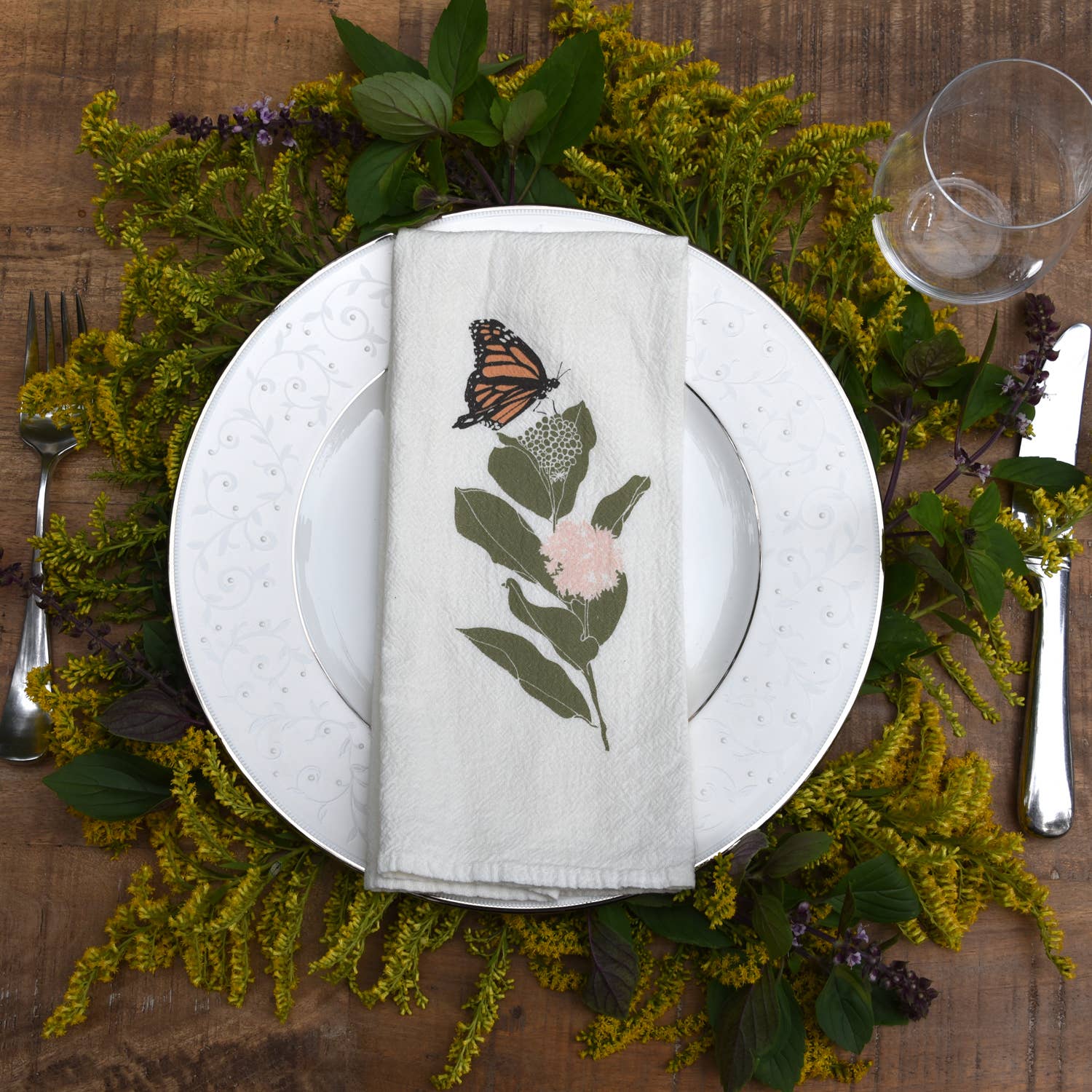 A charming white napkin towel with a playful bee and floral pattern is laid out next to a white plate crowned by yellow flora and leaves.