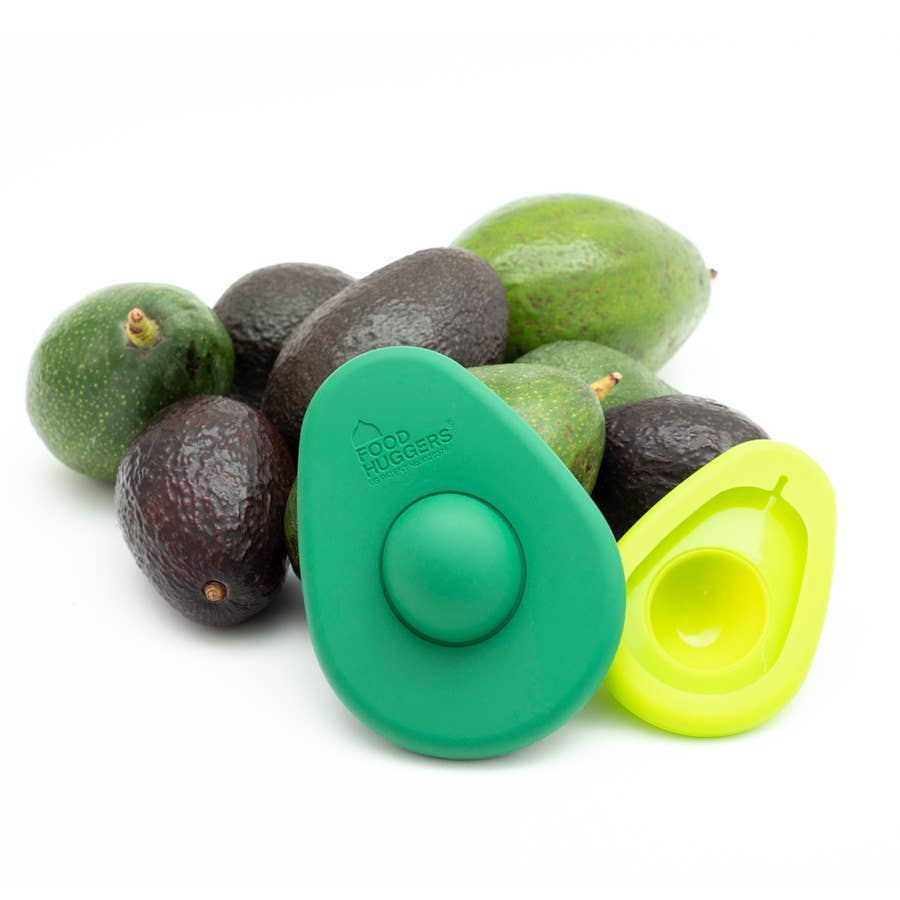 A pile of avocados in various shades of green and black sits on a white surface. In front of them are two Avocado Huggers, a large dark green one and a small light green one, demonstrating how they are perfectly shaped to keep avocados fresh.
