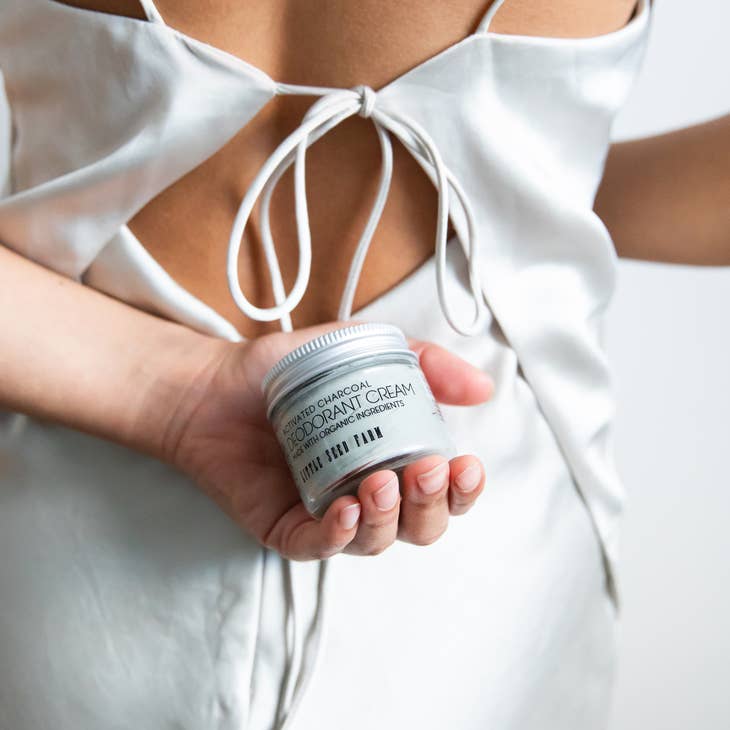 A woman in a white silk top holds a jar of Activated Charcoal Deodorant Cream from Little Seed Farm behind her back. The top has an open back with a bow tie detail.