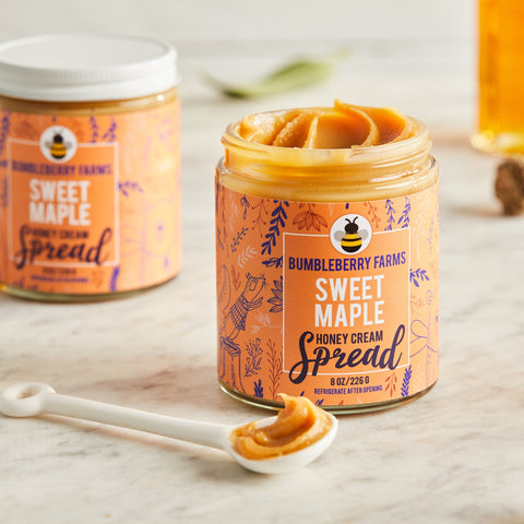 Two jars of Bumbleberry Farms Sweet Maple Honey Cream Spread are shown on a white marble surface. The jar in the foreground is open, revealing the creamy texture of the product with a spoonful of the spread resting on the surface next to it. A second, sealed jar is visible in the background.