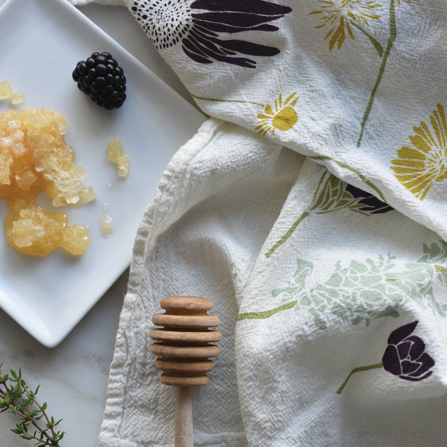 A white kitchen towel with a cute bee and floral pattern is laid out next to a white plate holding a glistening honeycomb and a fresh blackberry, with a wooden honey dipper and green sprig of thyme nearby.