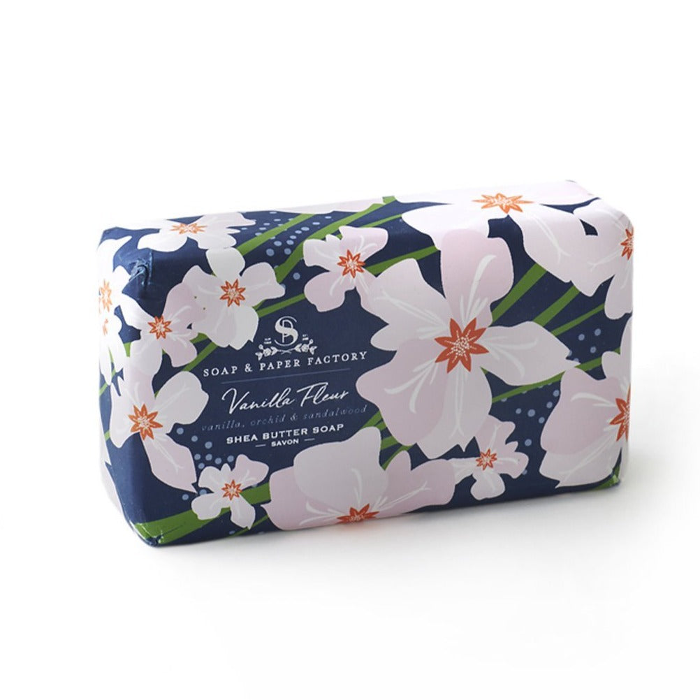 A "Vanilla Fleur" 'Soap & Paper Factory' shea butter soap bar with a dark blue wrapper. The wrapper features a pattern of large white flowers with light pink accents. The label lists scents of "vanilla, orchid & sandalwood."