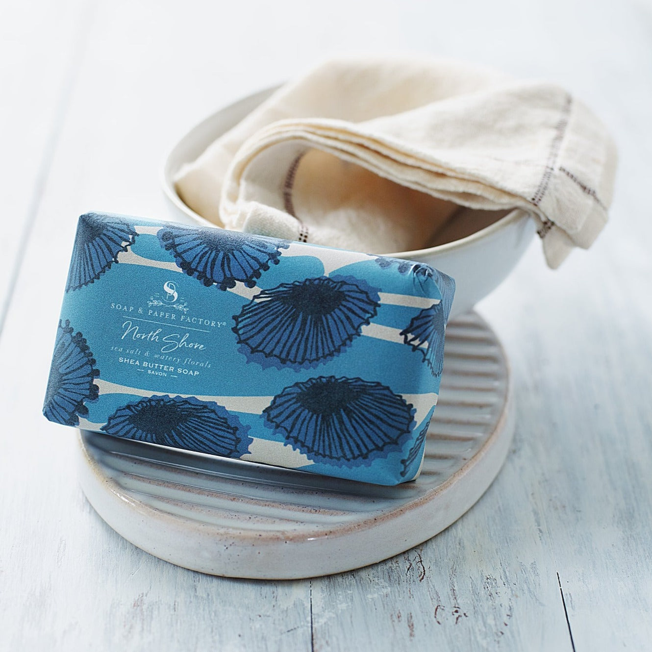 A blue-wrapped 'Soap & Paper Factory' shea butter soap bar sits on a ridged, oval soap dish. The wrapper, with a design of darker blue sea-shell-like flowers, is labeled "North Shore," with scents of "sea salt & watery florals."