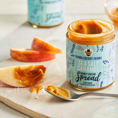 A jar of Bumbleberry Farms Sea Salt Caramel Honey Cream Spread is shown open on a white surface. A spoonful of the golden-brown spread rests on the surface, along with several slices of red apple, suggesting it can be used as a dip.