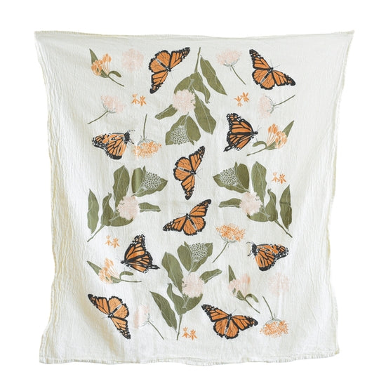 A lovely white cotton kitchen towel from, it is decorated with a playful pattern of monarch butterflies and delicate orange flowers with green leaves.