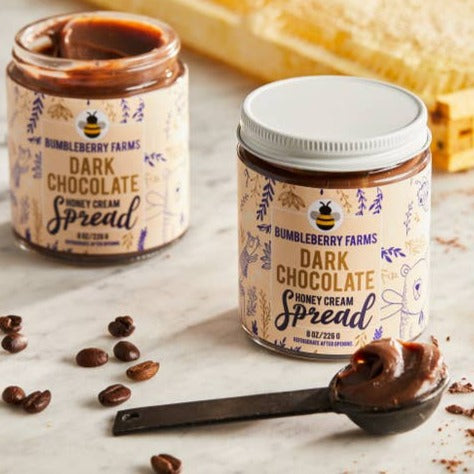 Two jars of Bumbleberry Farms Dark Chocolate Honey Cream Spread are featured, one open and revealing its tasty, rich texture and the other still sealed, promising an untouched indulgence. A spoonful of the luscious, dark spread invites for a delicious treat, while scattered coffee beans hint at a deep complementary flavor. The scene, with its warm, inviting tones, is designed to awaken a craving for this luxurious, sweet treat.