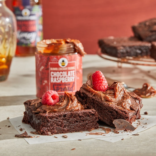Two delicious brownies topped with a swirl of chocolate spread and a fresh raspberry are shown on a white surface. A partially open jar of Bumbleberry Farms Chocolate Raspberry Honey Cream Spread is visible in the background, suggesting the source of the rich topping.