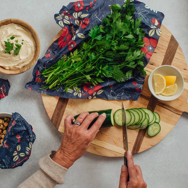 Hand slicing cucumber on a wooden board with fresh herbs on bee's wrap, lemon wedges, and hummus in a bowl nearby.