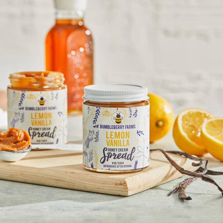 Two jars of Bumbleberry Farms Lemon Vanilla Honey Cream Spread are shown. The jar in the foreground is sealed, while a second jar behind it is open with a spoonful of the creamy spread next to it. Slices of fresh lemon and vanilla pods are scattered around the jars.