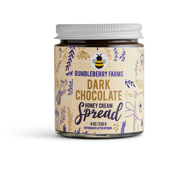 A jar of Bumbleberry Farms Dark Chocolate Honey Cream Spread is shown. The clear glass jar with a white lid holds a rich, dark-colored spread. The label is a light beige with a dark blue design of leaves and a prominent bee logo at the top. The product is identified as an 8 oz. honey cream spread.