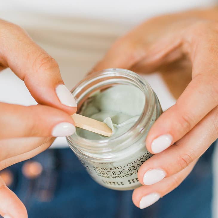 Hands with manicured nails use a small wooden stick to mix the gray Little Seed Farm Activated Charcoal deodorant cream inside its glass jar.