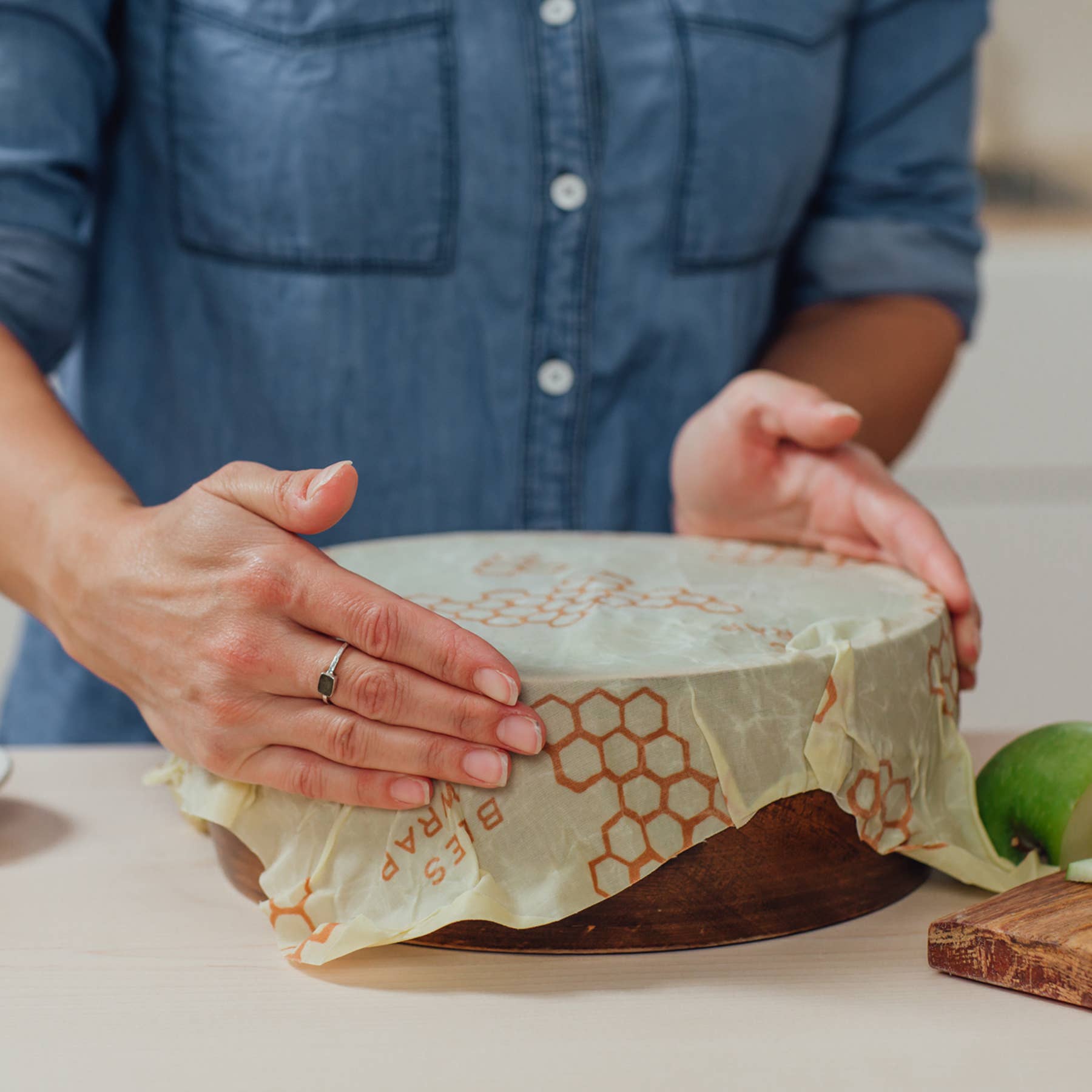 Person's hands covering a wooden bowl with a beeswax wrap printed with orange honeycomb patterns, on a kitchen counter.