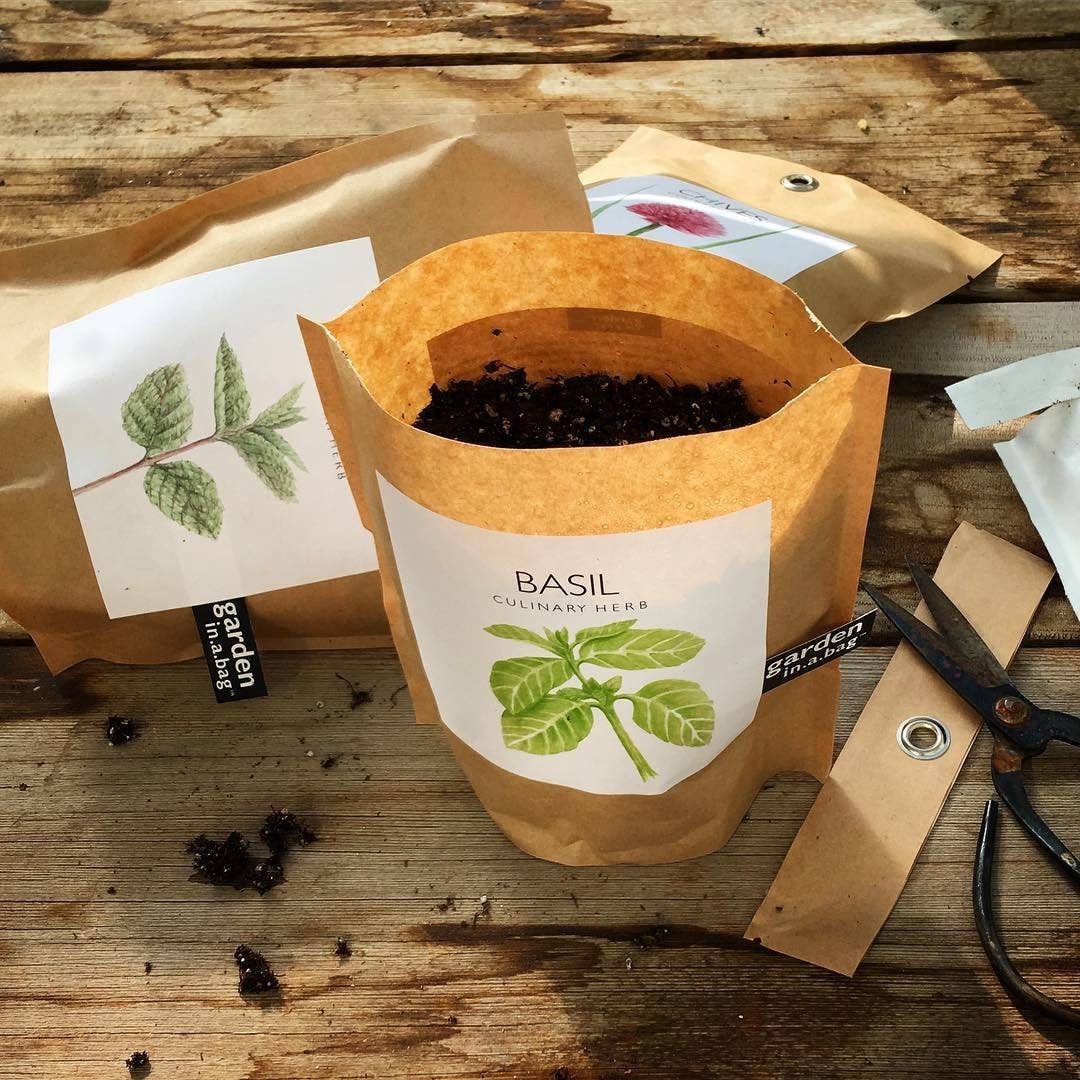 A brown paper "garden in a bag" pouch for "BASIL" is shown on a wooden table, partially filled with soil. Surrounding it are other pouches.