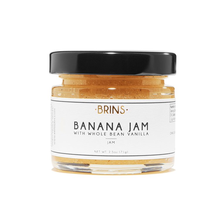 A small glass jar of "BRINS" Banana Jam with a black lid. The white label features the flavor and a description, "WITH WHOLE BEAN VANILLA" in black text. The golden yellow jam is visible inside the jar.