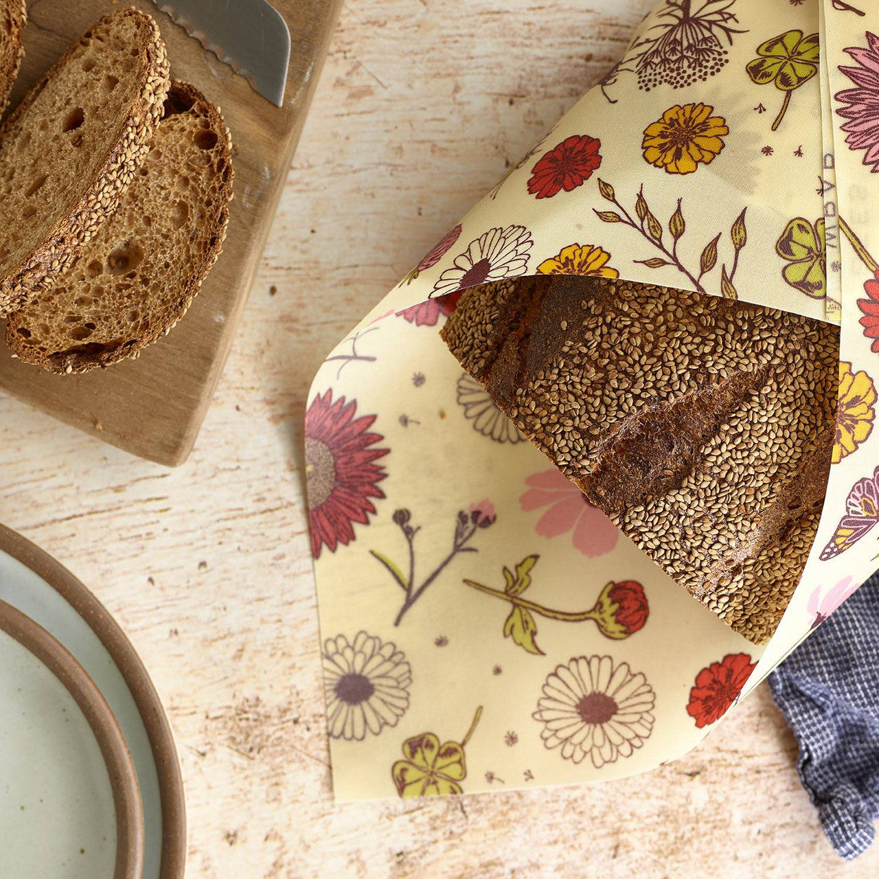 Close-up of a beeswax food wrap with floral print wrapped around a loaf of seeded bread on a wooden surface, with a plate and cloth napkin nearby.