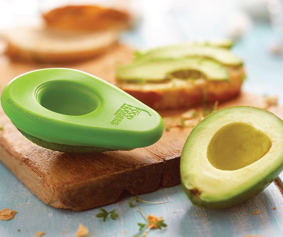 A delightful scene of sustainable food storage, where a reusable silicone avocado saver keeps a fresh avocado half from browning. A bright orange food saver is also shown covering a lime, all arranged with a bowl of guacamole and nachos.