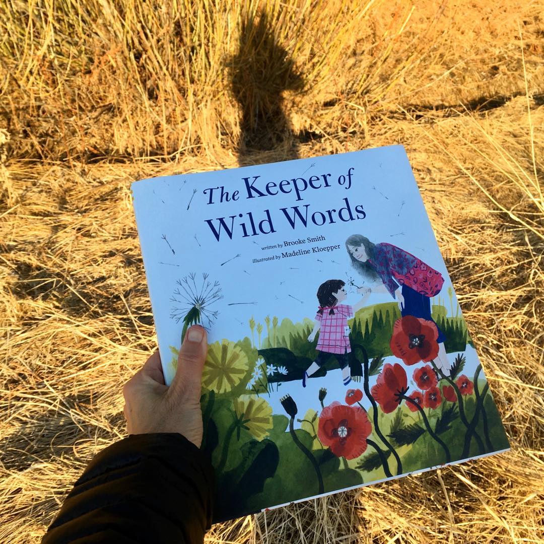 Close-up of the cover of the book "The Keeper of Wild Words" standing upright on a yellow grass surface.