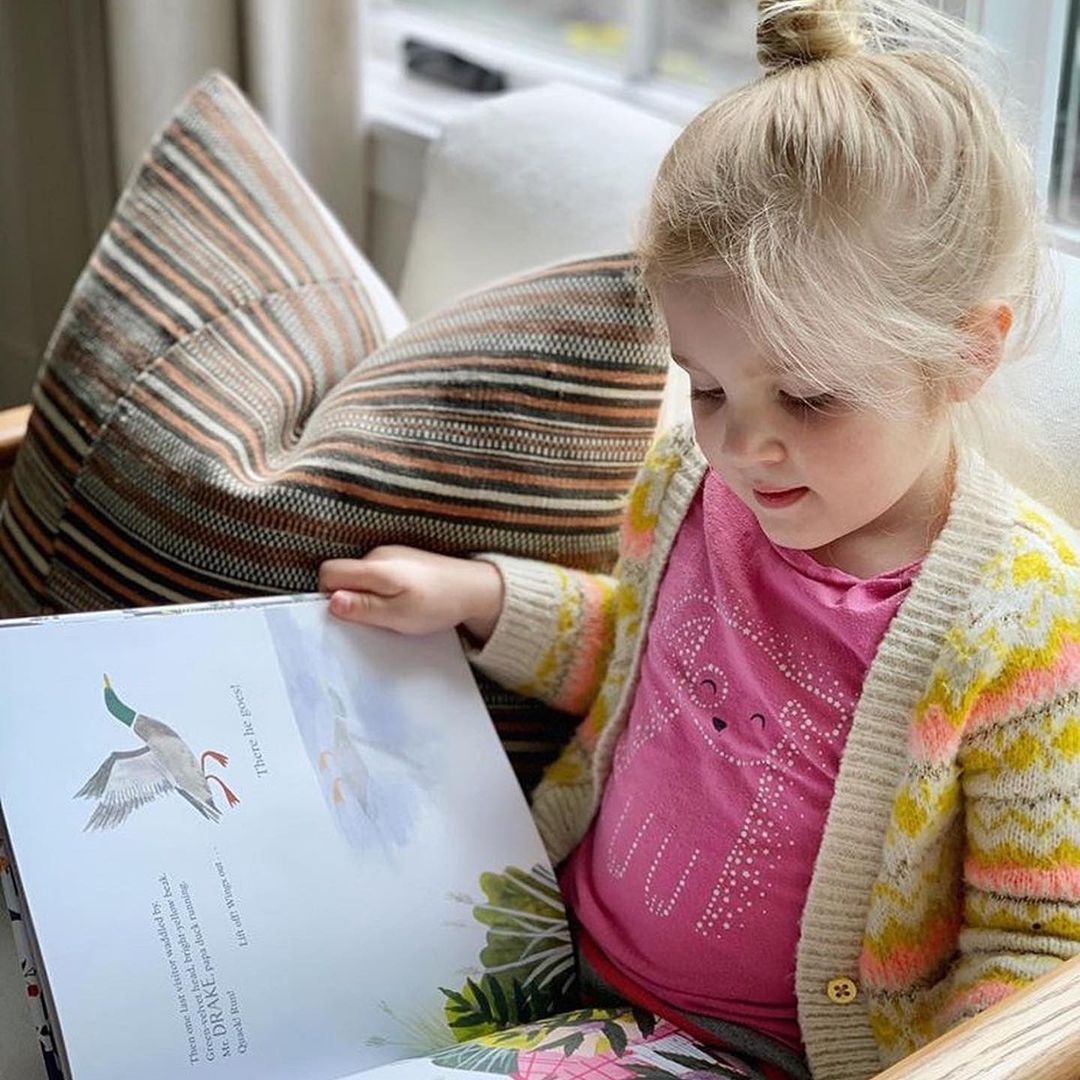 Blonde girl sitting on a couch, reading the hardcover book "The Keeper of Wild Words" with colorful nature illustrations visible on the open pages.
