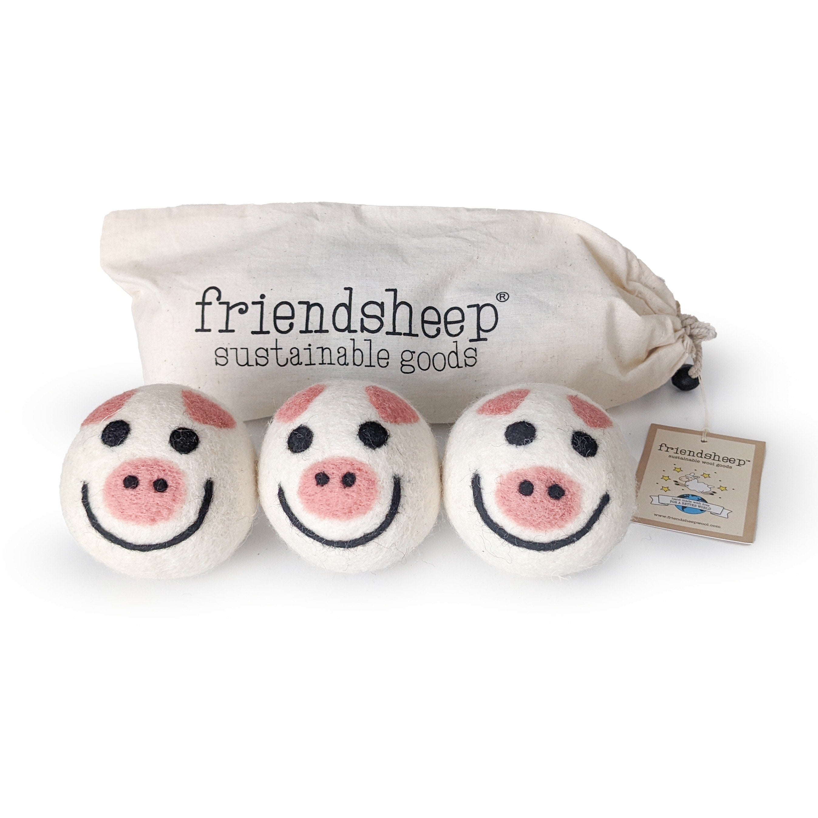 A set of three Friendsheep sustainable goods wool dryer balls are shown, each decorated with a cute pig face. They are sitting in front of their white canvas drawstring bag.